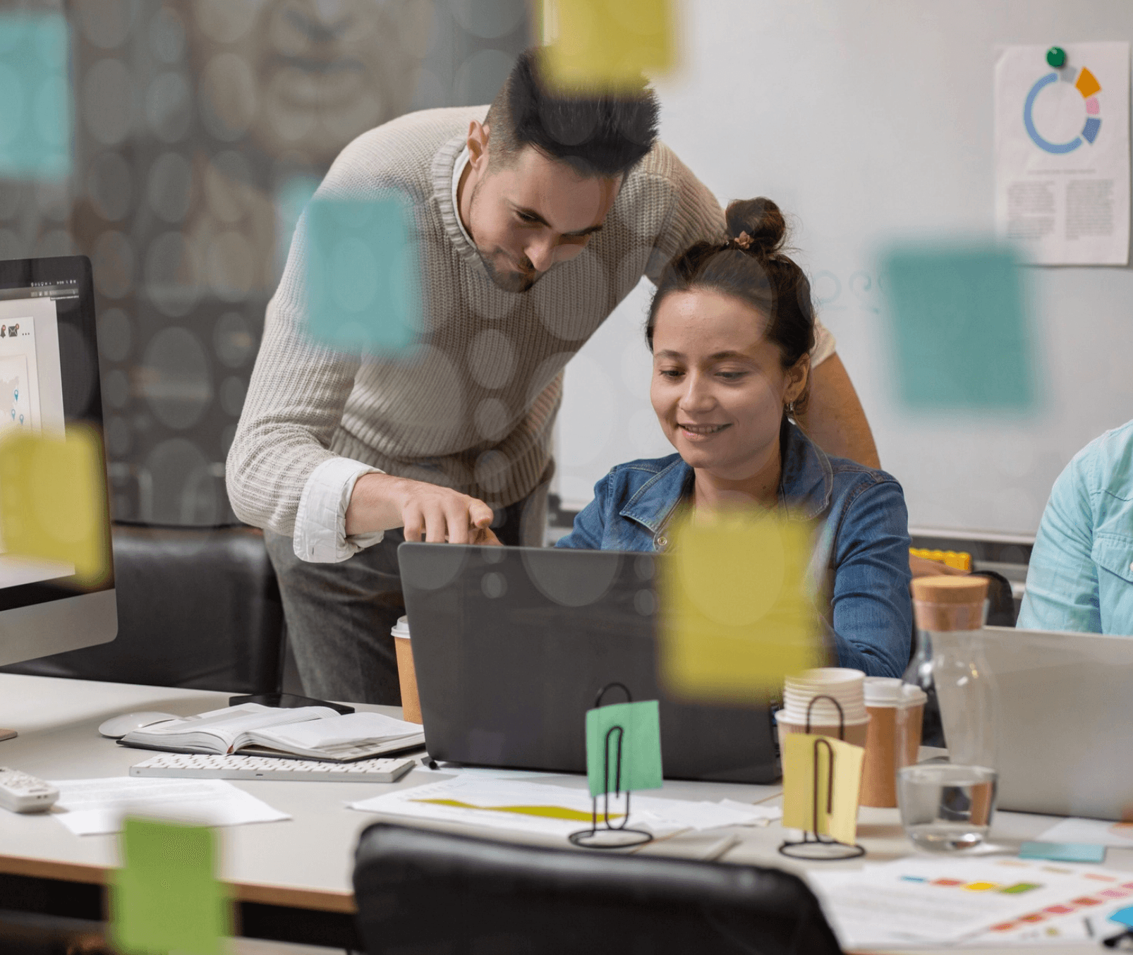 Two colleagues reviewing work on a laptop in an office, seen through a glass wall covered in sticky notes